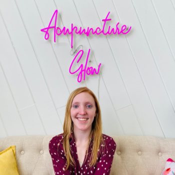 Portrait of receptionist, Leanna, sitting on the couch in the reception area of Seneca Falls Acupuncture under the pink "Acupuncture Glow" neon sign.