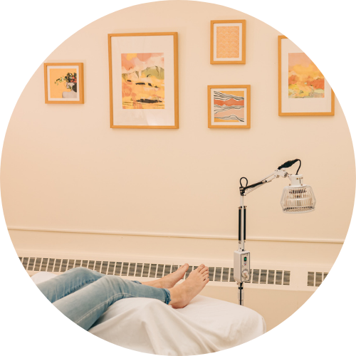 View of patient's feet as they rest on a treatment table with a heat lamp and yellow art in the background at Seneca Falls Acupuncture.