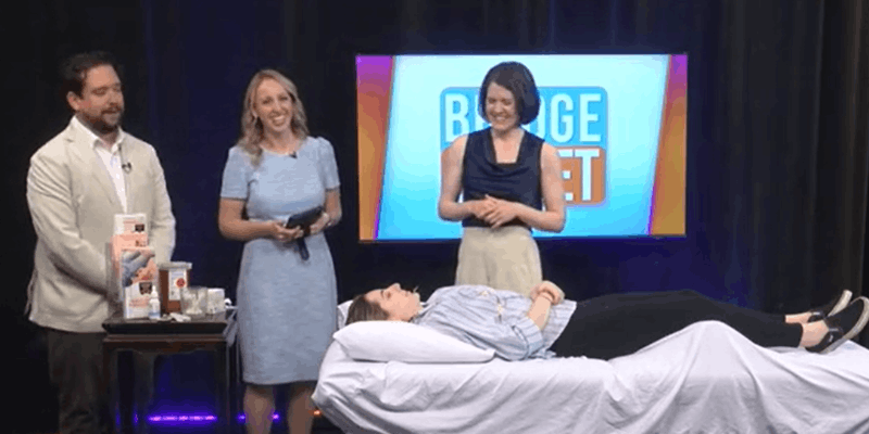 Image of acupuncturist Michelle Grasek speaking with Bridge Street hosts on set while volunteer rests with acupuncture on a treatment table in front of them.